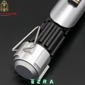 May include: A silver and black lightsaber hilt with a ribbed black center section and a silver handle. The lightsaber has a silver metal loop and the name "EZRA" in green digital font. The logo "ASTROMX SABERS" is in the upper left corner.