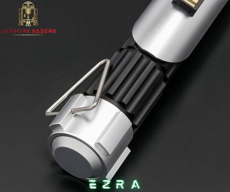 May include: A silver and black lightsaber hilt with a ribbed black center section and a silver handle. The lightsaber has a silver metal loop and the name "EZRA" in green digital font. The logo "ASTROMX SABERS" is in the upper left corner.