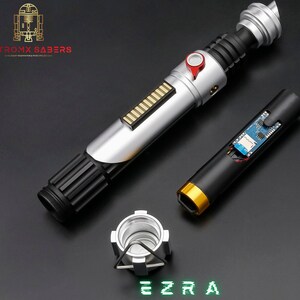 May include: A disassembled lightsaber with silver, black, and red accents, featuring a detailed hilt and exposed internal components. The text "EZRA" is displayed in green, along with the "ASTROMX SABERS" logo.