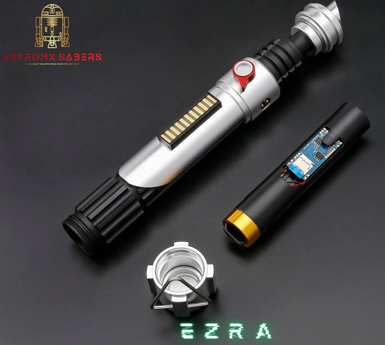 May include: A disassembled lightsaber with silver, black, and red accents, featuring a detailed hilt and exposed internal components. The text "EZRA" is displayed in green, along with the "ASTROMX SABERS" logo.