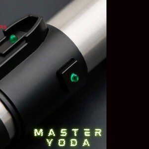 May include: A lightsaber with a black handle and silver details. It features two green lights and the text "MASTER YODA" in green. The "ASTRO SABERS" logo is visible in the top left corner.