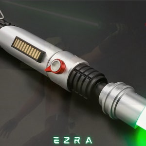 May include: A silver and black lightsaber with a green blade. The hilt features a red button and a gold and black rectangular detail. The word "EZRA" is displayed in green block letters at the bottom. The Astromx Sabers logo is in the upper right corner.