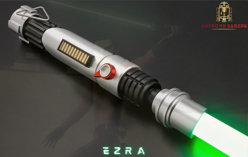 May include: A silver and black lightsaber with a green blade. The hilt features a red button and a gold and black rectangular detail. The word "EZRA" is displayed in green block letters at the bottom. The Astromx Sabers logo is in the upper right corner.