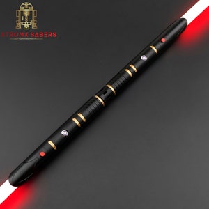 K&ouml;nnte beinhalten: Schwarzes Lichtschwert mit goldenen Akzenten und roter Klinge. Das Lichtschwert hat mehrere Kn&ouml;pfe und ein schlankes Design. Das Logo "ASTROMX SABERS" ist oben links sichtbar.
