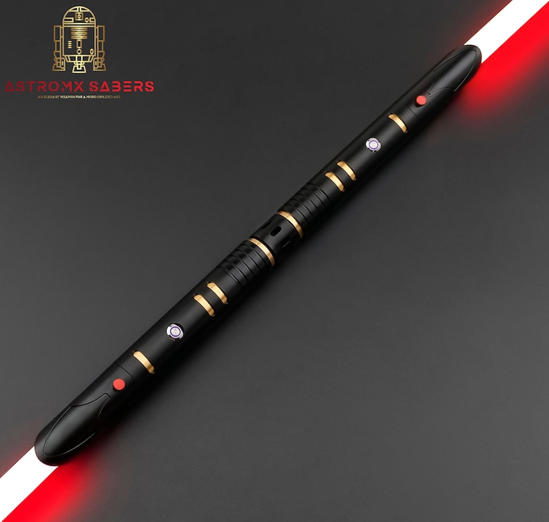 K&ouml;nnte beinhalten: Schwarzes Lichtschwert mit goldenen Akzenten und roter Klinge. Das Lichtschwert hat mehrere Kn&ouml;pfe und ein schlankes Design. Das Logo "ASTROMX SABERS" ist oben links sichtbar.