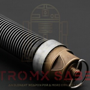 Puede incluir: Primer plano de una empu&ntilde;adura de sable l&aacute;ser plateada y bronce con un dise&ntilde;o acanalado. La empu&ntilde;adura tiene un peque&ntilde;o anillo en el extremo y el texto "ASTROMX SABERS" es visible en la parte inferior de la imagen.  "AN ELEGANT WEAPON FOR A MORE CIVILIZED AGE" tambi&eacute;n es visible en la parte inferior de la imagen.