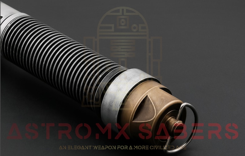 Puede incluir: Primer plano de una empu&ntilde;adura de sable l&aacute;ser plateada y bronce con un dise&ntilde;o acanalado. La empu&ntilde;adura tiene un peque&ntilde;o anillo en el extremo y el texto "ASTROMX SABERS" es visible en la parte inferior de la imagen.  "AN ELEGANT WEAPON FOR A MORE CIVILIZED AGE" tambi&eacute;n es visible en la parte inferior de la imagen.