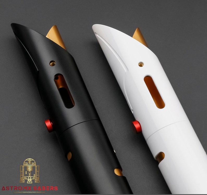 K&ouml;nnte beinhalten: Zwei Lichtschwertgriffe, einer schwarz und einer wei&szlig;, mit goldenen Akzenten und roten Kn&ouml;pfen. Die Griffe haben ein schlankes, modernes Design. Das Logo "Astromx Sabers" ist unten sichtbar.