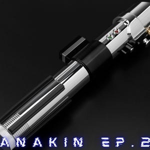 May include: A metallic silver and black lightsaber with the text "ANAKIN EP. 2" in blue. The lightsaber has a detailed design with buttons and a control panel. The logo "ANTROX SABERS" is in the upper left corner.
