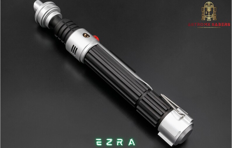 May include: A detailed lightsaber prop with a black ribbed handle, silver accents, and a red button. The text "EZRA" is displayed in green, pixelated font. The logo for "ASTROMX SABERS" is in the upper right corner.