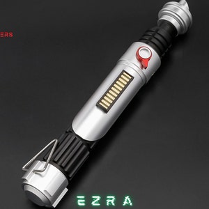 May include: A silver and black lightsaber with a red button and a series of gold bars. The text "EZRA" is displayed in green. The logo "ASTROMX SABERS" is in the upper left corner.