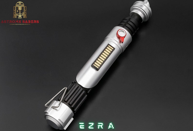 May include: A silver and black lightsaber with a red button and a series of gold bars. The text "EZRA" is displayed in green. The logo "ASTROMX SABERS" is in the upper left corner.