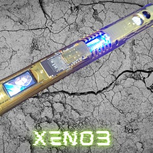 May include: A gold-colored lightsaber with blue and white internal lighting. The lightsaber has a small screen displaying an image and the text "XENO3" in green. The lightsaber is on a cracked gray surface.