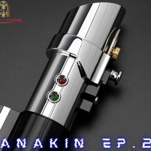 May include: A close-up of a metallic lightsaber hilt with a polished chrome finish. The hilt features red and green activation buttons, a gold-colored accent, and the text "ANAKIN EP. 2" in blue. The logo "ASTROMA SABERS" is in the upper left corner.