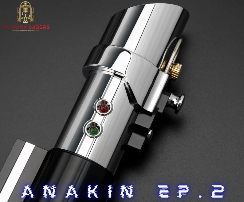 May include: A close-up of a metallic lightsaber hilt with a polished chrome finish. The hilt features red and green activation buttons, a gold-colored accent, and the text "ANAKIN EP. 2" in blue. The logo "ASTROMA SABERS" is in the upper left corner.