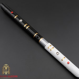 K&ouml;nnte beinhalten: Ein schwarz-wei&szlig;es Lichtschwert mit goldenen und silbernen Akzenten. Der Griff hat einen roten Knopf und goldfarbene Ringe. Das Lichtschwert tr&auml;gt unten den Text "ASTRO MX SABERS".