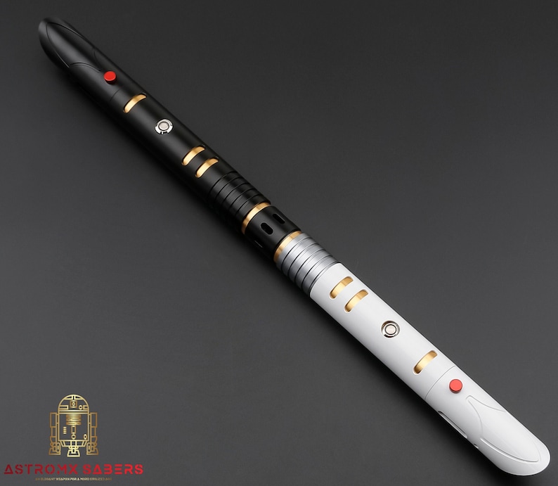K&ouml;nnte beinhalten: Ein schwarz-wei&szlig;es Lichtschwert mit goldenen und silbernen Akzenten. Der Griff hat einen roten Knopf und goldfarbene Ringe. Das Lichtschwert tr&auml;gt unten den Text "ASTRO MX SABERS".