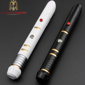 K&ouml;nnte beinhalten: Zwei Lichtschwerter, eines wei&szlig; und eines schwarz, mit goldenen und silbernen Akzenten. Jedes hat einen roten Knopf und ein zylindrisches Design. Der Text "ASTROMX SABERS" ist oben links sichtbar.