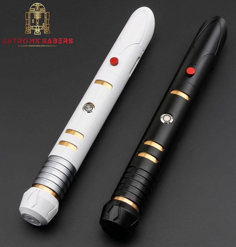 K&ouml;nnte beinhalten: Zwei Lichtschwerter, eines wei&szlig; und eines schwarz, mit goldenen und silbernen Akzenten. Jedes hat einen roten Knopf und ein zylindrisches Design. Der Text "ASTROMX SABERS" ist oben links sichtbar.