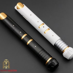 K&ouml;nnte beinhalten: Zwei futuristische Lichtschwerter, eines schwarz und eines wei&szlig;, mit goldenen und silbernen Akzenten. Das schwarze Lichtschwert hat einen schwarzen Griff und eine goldene Spitze. Das wei&szlig;e Lichtschwert hat einen silbernen Griff und eine goldene Spitze. Die Schwerter liegen auf einer dunkelgrauen Oberfl&auml;che.
