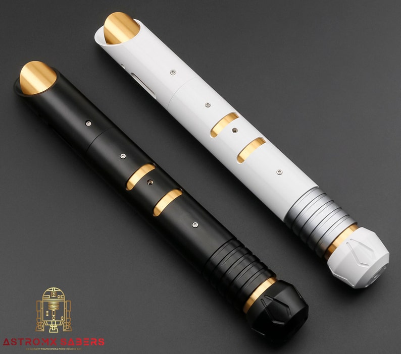 K&ouml;nnte beinhalten: Zwei futuristische Lichtschwerter, eines schwarz und eines wei&szlig;, mit goldenen und silbernen Akzenten. Das schwarze Lichtschwert hat einen schwarzen Griff und eine goldene Spitze. Das wei&szlig;e Lichtschwert hat einen silbernen Griff und eine goldene Spitze. Die Schwerter liegen auf einer dunkelgrauen Oberfl&auml;che.