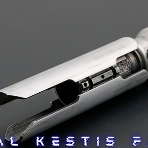 K&ouml;nnte beinhalten: Ein silberner Lichtschwertgriff mit einem schwarzen Griff und einer silbernen Klinge. Der Griff hat eine Reihe kleiner, runder L&ouml;cher und einen schwarzen Knopf an der Seite. Der Text "CAL KESTIS F/O" ist am unteren Rand des Bildes sichtbar.