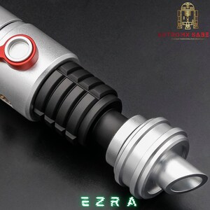 May include: A silver and black lightsaber with red accents. The lightsaber has a ribbed black grip and a silver emitter. The word "EZRA" is displayed in green block letters at the bottom of the image. The logo "ANTROMX SABERS" is in the upper right corner.