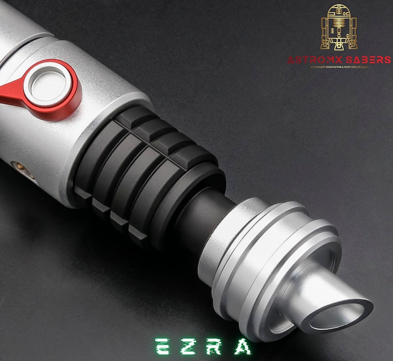 May include: A silver and black lightsaber with red accents. The lightsaber has a ribbed black grip and a silver emitter. The word "EZRA" is displayed in green block letters at the bottom of the image. The logo "ANTROMX SABERS" is in the upper right corner.