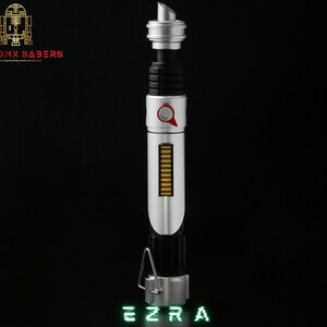 May include: A silver and black lightsaber with a red button and a series of gold bars. The text "EZRA" is displayed in green at the bottom. The logo "ASTROMX SABERS" is in the upper left corner.