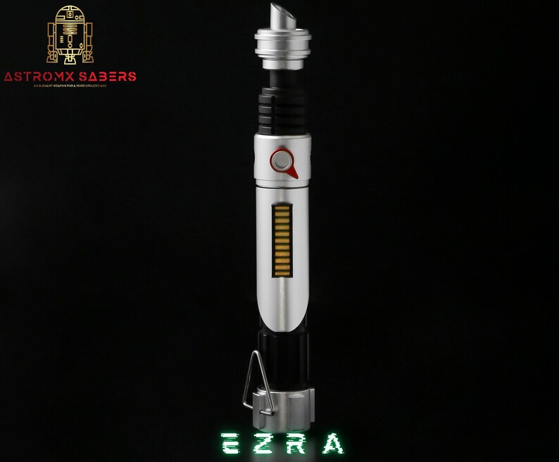 May include: A silver and black lightsaber with a red button and a series of gold bars. The text "EZRA" is displayed in green at the bottom. The logo "ASTROMX SABERS" is in the upper left corner.