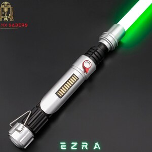 May include: A silver and black lightsaber with a bright green blade. The hilt features a red button and a rectangular panel with gold-colored bars. The text "EZRA" is displayed in green, pixelated font. The logo "ASTROMX SABERS" is in the upper left corner.