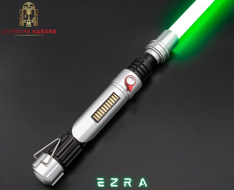 May include: A silver and black lightsaber with a bright green blade. The hilt features a red button and a rectangular panel with gold-colored bars. The text "EZRA" is displayed in green, pixelated font. The logo "ASTROMX SABERS" is in the upper left corner.