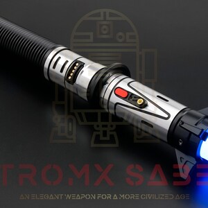 Puede incluir: Un sable de luz plateado y negro con una hoja azul. El sable de luz tiene un agarre texturizado y un bot&oacute;n rojo en el lateral. El texto "ASTROMX SABERS" es visible en la esquina inferior derecha de la imagen.