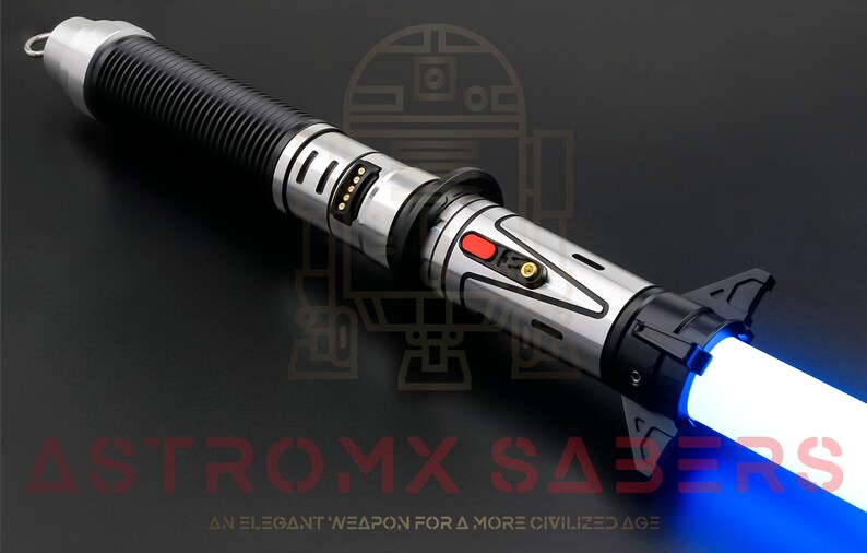 Puede incluir: Un sable de luz plateado y negro con una hoja azul. El sable de luz tiene un agarre texturizado y un bot&oacute;n rojo en el lateral. El texto "ASTROMX SABERS" es visible en la esquina inferior derecha de la imagen.