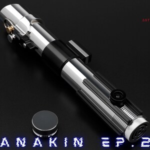 May include: A silver and black lightsaber with a cylindrical design, featuring intricate details and a button. A silver cap sits beside the lightsaber. The text "ANAKIN EP.2" is displayed in blue at the bottom.