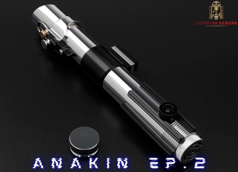 May include: A silver and black lightsaber with a cylindrical design, featuring intricate details and a button. A silver cap sits beside the lightsaber. The text "ANAKIN EP.2" is displayed in blue at the bottom.