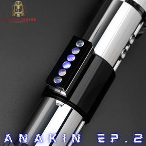May include: A close-up of a lightsaber with a black and silver design. The lightsaber has a black rectangular control panel with six illuminated circular lights. The text "ANAKIN EP. 2" is displayed in blue at the bottom.