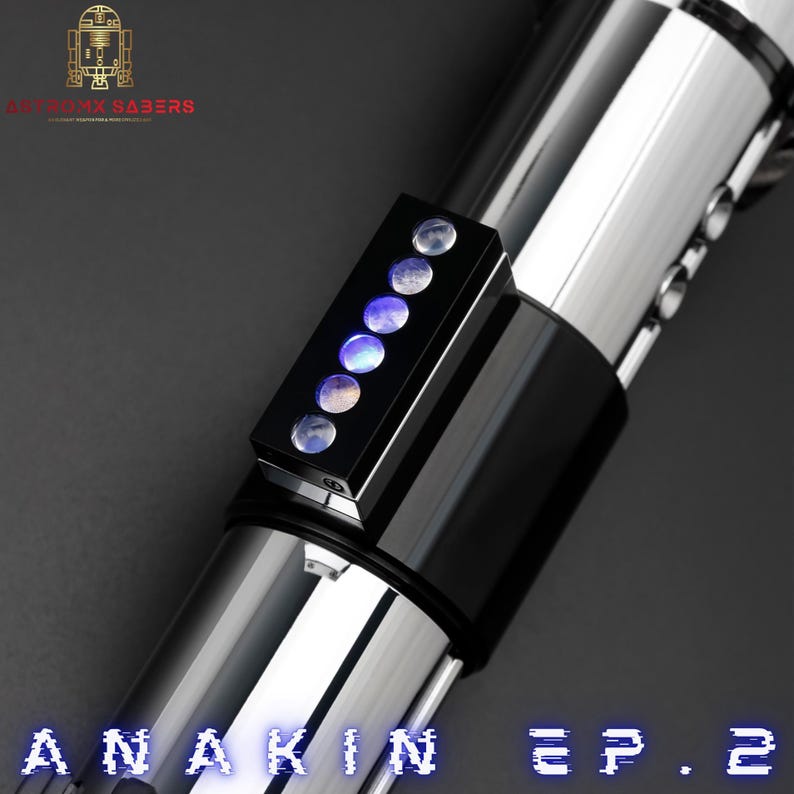 May include: A close-up of a lightsaber with a black and silver design. The lightsaber has a black rectangular control panel with six illuminated circular lights. The text "ANAKIN EP. 2" is displayed in blue at the bottom.