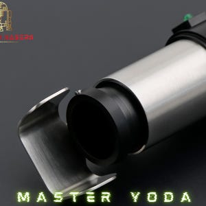 May include: A lightsaber hilt with a silver and black design is shown in close-up. The lightsaber has a cylindrical body with a silver finish and a black grip. The text "MASTER YODA" is displayed in green at the bottom.