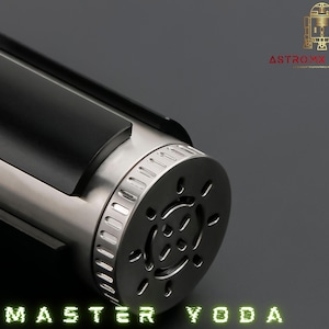 May include: A close-up of a silver and black lightsaber hilt. The hilt has a cylindrical design with a black grip and a detailed end cap. The text "MASTER YODA" is displayed in green, stylized lettering.