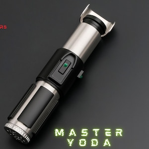 May include: A silver and black lightsaber with the text "MASTER YODA" in green neon lettering. The lightsaber features a silver hilt, black grip, and a green button. The Astromx Sabers logo is in the upper left corner.