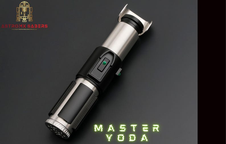 May include: A silver and black lightsaber with the text "MASTER YODA" in green neon lettering. The lightsaber features a silver hilt, black grip, and a green button. The Astromx Sabers logo is in the upper left corner.