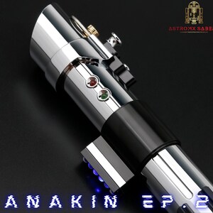 May include: A silver and black lightsaber with red and green buttons, and the text "ANAKIN EP. 2" in blue. The lightsaber has a metallic finish and is angled against a dark background. The logo "ASTROMX SABERS" is in the upper right corner.