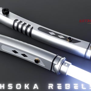 May include: Two silver lightsabers with white blades are displayed against a dark background. The hilts have black and gold accents, with the text "AHSOKA REBELS" at the bottom. The top right corner features a gold logo and the text "ASTROMX SABERS".
