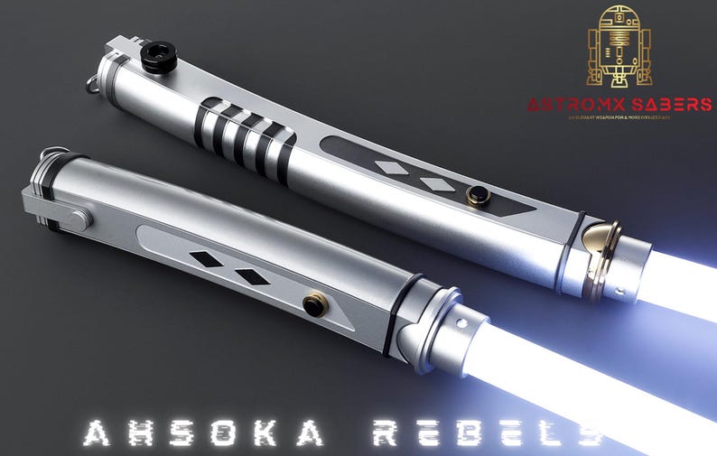 May include: Two silver lightsabers with white blades are displayed against a dark background. The hilts have black and gold accents, with the text "AHSOKA REBELS" at the bottom. The top right corner features a gold logo and the text "ASTROMX SABERS".