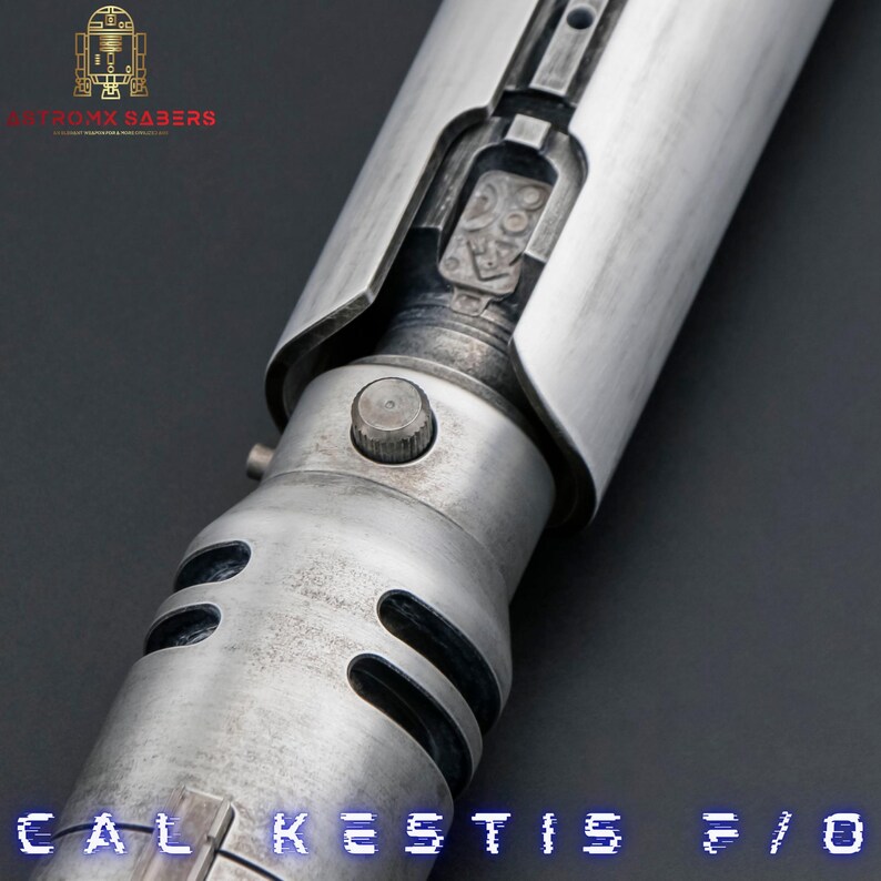 K&ouml;nnte beinhalten: Nahaufnahme einer silbernen Lichtschwert-Griff mit einem schwarzen Knopf und einem silbernen und schwarzen Design. Der Griff hat eine verwitterte Optik und der Text "CAL KESTIS F/O" ist unten sichtbar.