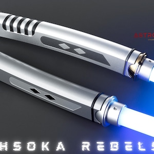 May include: Two silver lightsabers with white and blue blades are displayed against a dark background. The lightsabers have detailed handles with black accents and diamond-shaped designs. The text "AHSOKA REBELS" is at the bottom.