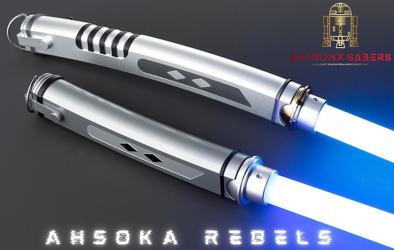 May include: Two silver lightsabers with white and blue blades are displayed against a dark background. The lightsabers have detailed handles with black accents and diamond-shaped designs. The text "AHSOKA REBELS" is at the bottom.