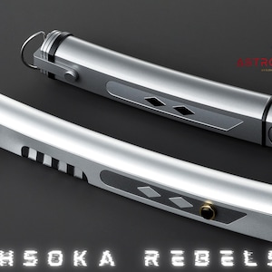 May include: Two silver lightsabers with black and gold accents, featuring a diamond pattern and ring details. The text "AHSOKA REBELS" is displayed at the bottom, with the logo "ASTROMX SABERS" in the upper right corner.