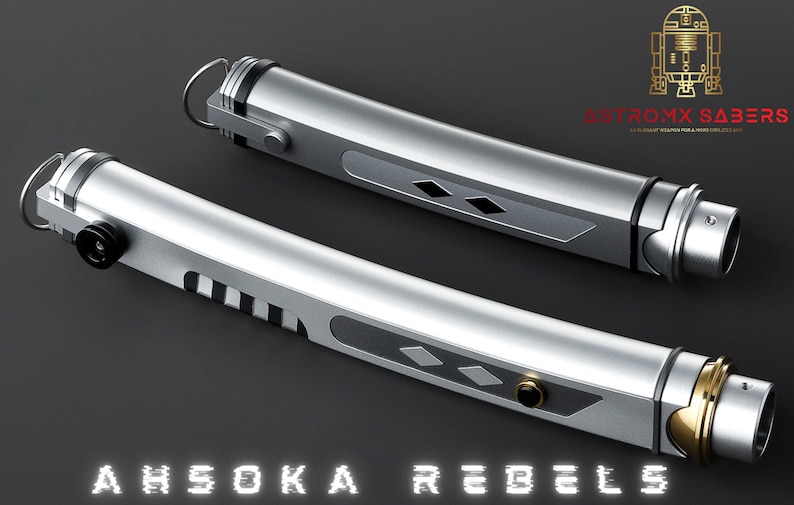 May include: Two silver lightsabers with black and gold accents, featuring a diamond pattern and ring details. The text "AHSOKA REBELS" is displayed at the bottom, with the logo "ASTROMX SABERS" in the upper right corner.